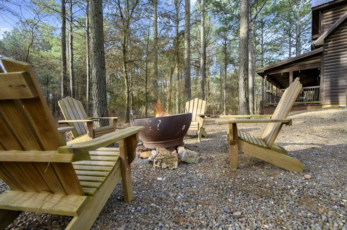 Fire pit with four Adirondack chairs on the gravel behind Livin' the Dream, the cabin and its deck behind