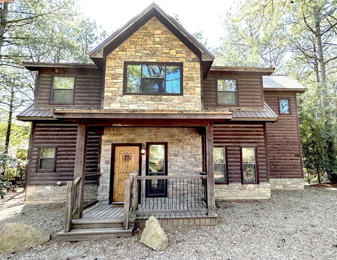 Front of Livin' the Dream, a two-story log cabin with a stone gable and a covered front porch among pine trees