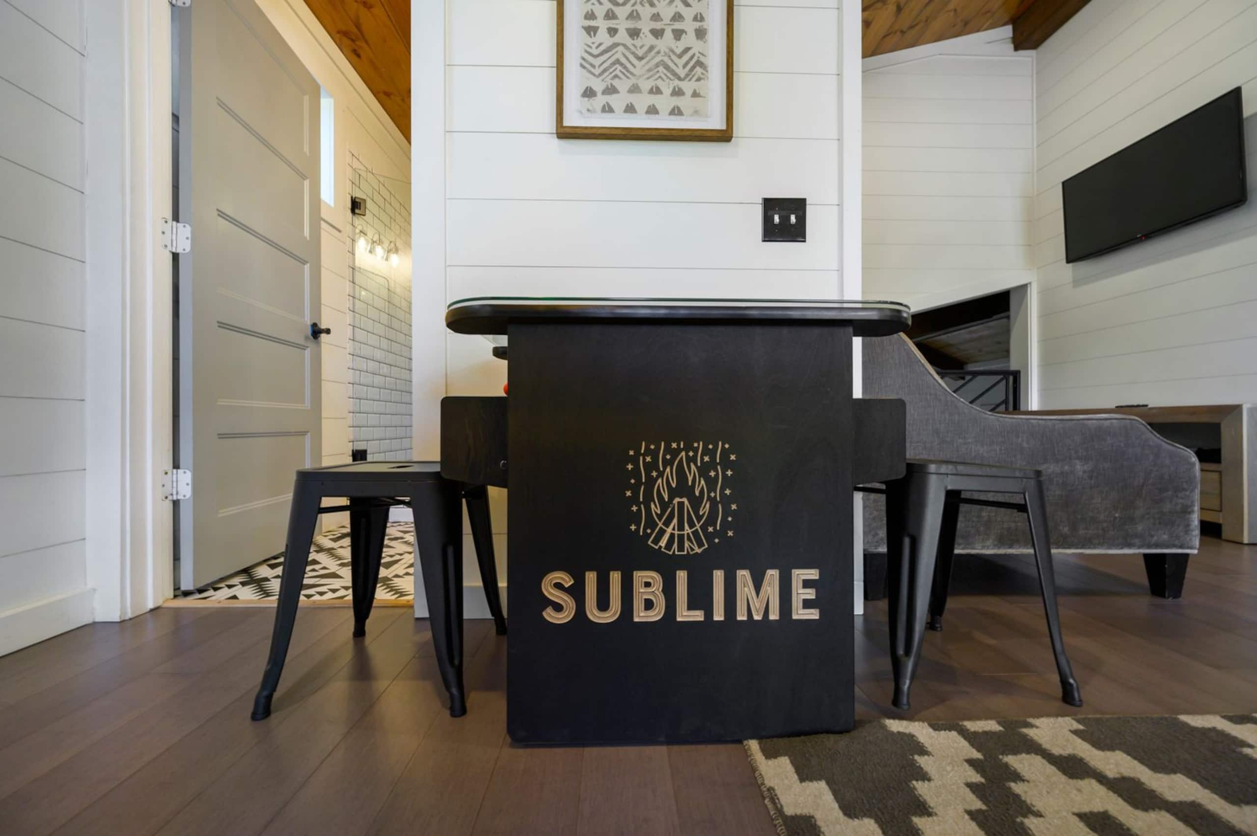Tabletop arcade at Sublime Retreat, its side lettered SUBLIME with a campfire logo, with stools on either side